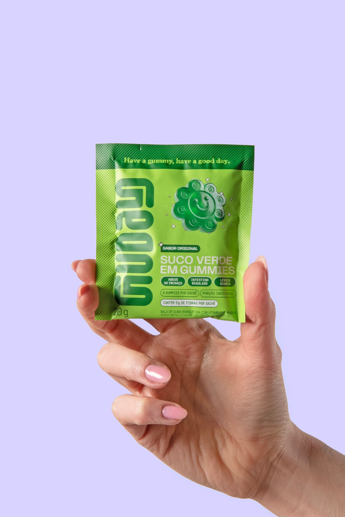 Mão segurando o sachê de Suco Verde em Gummies Guday sobre fundo lilás