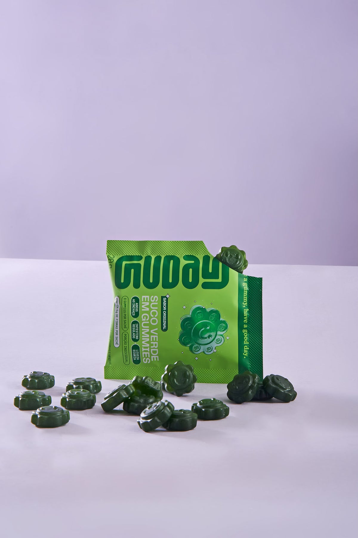 Sachê de Suco Verde em Gummies Guday rasgado, com uma gummy saindo e outras espalhadas sobre fundo lilás